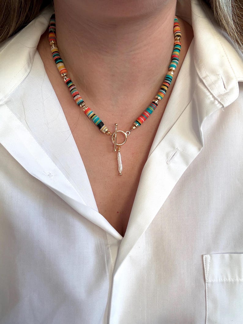 May include: A colorful beaded necklace with a gold clasp and a dangling charm. The necklace features a variety of colors, including red, orange, yellow, green, and blue. The clasp is a gold toggle style. The charm is a small, white, elongated shape.