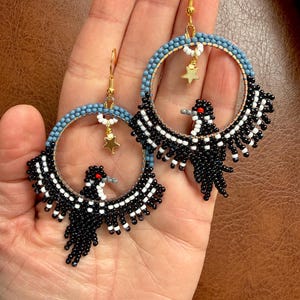 K&ouml;nnte beinhalten: Handgefertigte Perlen-Creolen mit einem schwarz-wei&szlig;en Vogeldesign und roten Akzenten. Die Reifen haben einen blau-goldenen Perlenrand mit einem goldenen Sternanh&auml;nger. Die Ohrringe werden auf einer Hand pr&auml;sentiert.