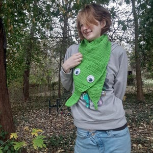 May include: A bright green, crocheted scarf with large, googly eyes. The scarf is designed to resemble a creature. The person is wearing a gray sweatshirt and blue jeans. The background is a natural outdoor setting with trees and fallen leaves.