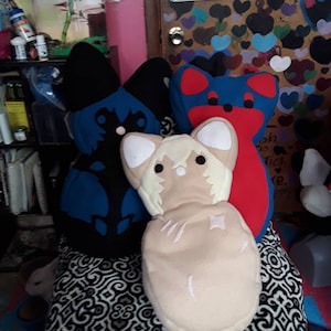 May include: Three plush toys in the shape of stylized animals. One is tan with white accents and stitched details. The other two are blue and red with black accents. They are arranged on a patterned surface.
