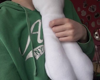 May include: A handmade white plush doll with button eyes. The doll has a long body and two leg-like appendages. The doll is being held by a person wearing a green hoodie.