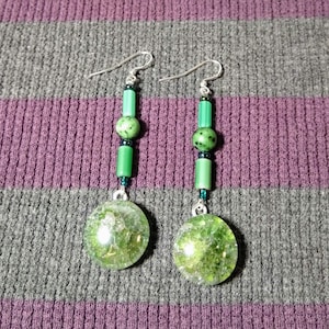 May include: A pair of green and silver earrings with a round green stone at the bottom. The earrings are made with small green beads and silver wire.