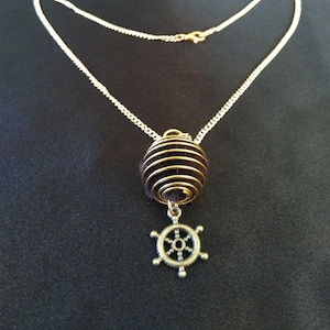 Könnte beinhalten: Eine silberfarbene Halskette mit einem goldfarbenen Spiralkäfig-Anhänger, der eine dunkelbraune Kugel umgibt. Ein silberfarbener Steuerrad-Anhänger hängt darunter. Die Halskette befindet sich auf einem schwarzen Hintergrund.