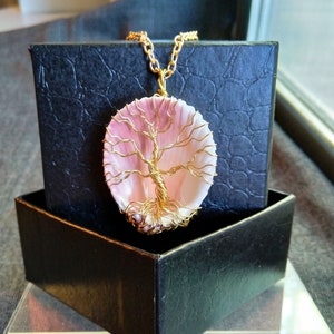 Könnte beinhalten: Ein goldener Draht-Lebensbaum-Anhänger in einer rosa Muschel, auf einer schwarzen Samtbox. Der Anhänger ist an einer goldenen Kette.
