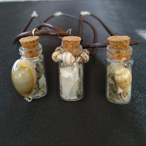 May include: Three small glass bottles with cork tops and brown leather cord necklaces. Each bottle contains seashells and other beach-themed items.