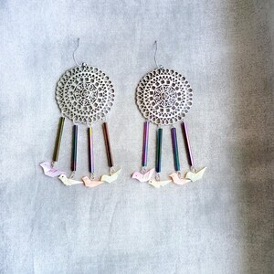 May include: Silver filigree earrings with dangling iridescent rods and small bird charms in pastel colors. The earrings have a circular design with intricate cut-out patterns and are suspended from silver hooks.