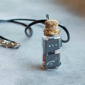May include: A small glass bottle necklace with a cork stopper. The bottle contains a small black circuit board with a phone icon and three dots.