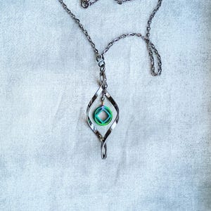 Può includere: Collana color argento con un ciondolo unico. Il ciondolo presenta un design attorcigliato e allungato con un piccolo ciondolo circolare nei toni del verde e del blu. La collana è esposta su uno sfondo bianco strutturato.