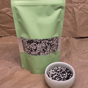 May include: A light green stand-up pouch with a clear rectangular window revealing a mixture of small black and white granular stones. Next to the pouch sits a small white ceramic bowl filled with the same black and white stone mixture.