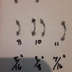 May include: Five silver body jewelry pieces with different styles and sizes. The jewelry pieces are arranged in three rows. The top row has two pieces with a ball on one end and a curved shape on the other. The middle row has three pieces with a pointed end on one side and a curved shape on the other. The bottom row has three numbers, 8, 10, and 11, and three fractions, 5/16, 3/8, and 7/16.