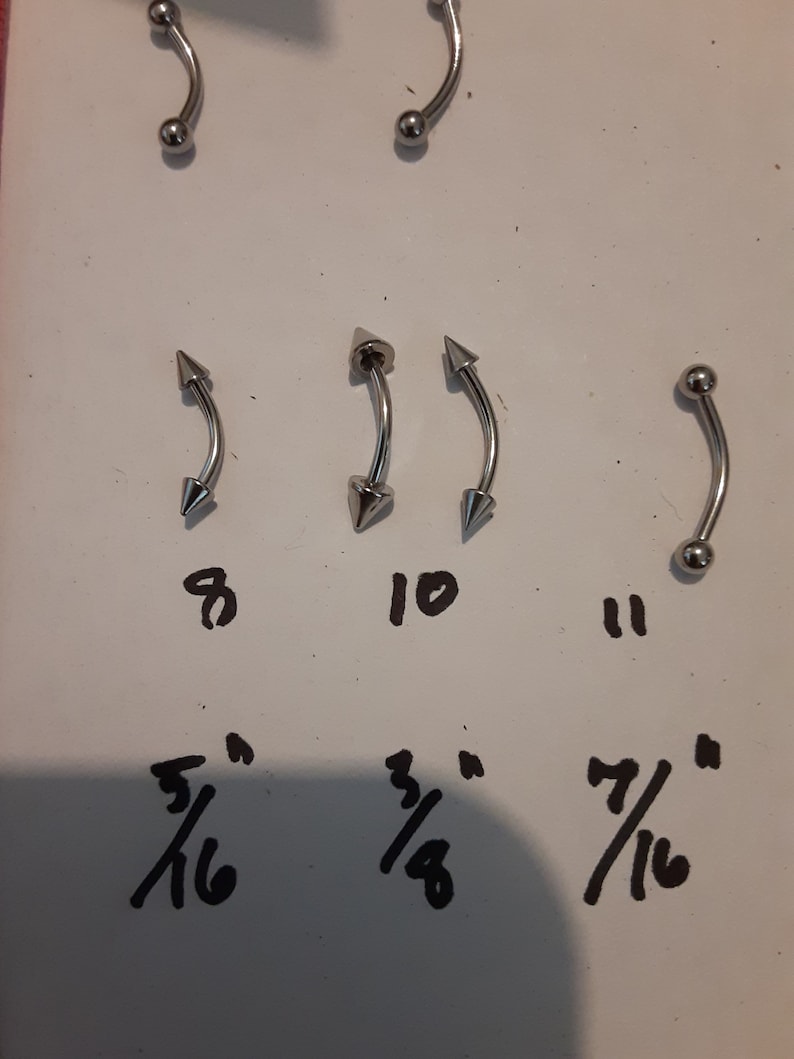 May include: Five silver body jewelry pieces with different styles and sizes. The jewelry pieces are arranged in three rows. The top row has two pieces with a ball on one end and a curved shape on the other. The middle row has three pieces with a pointed end on one side and a curved shape on the other. The bottom row has three numbers, 8, 10, and 11, and three fractions, 5/16, 3/8, and 7/16.