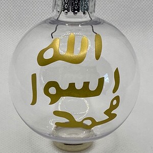 May include: Clear glass Christmas ornament with a gold Arabic calligraphy design that reads "Allah" and "Muhammad" in gold lettering.