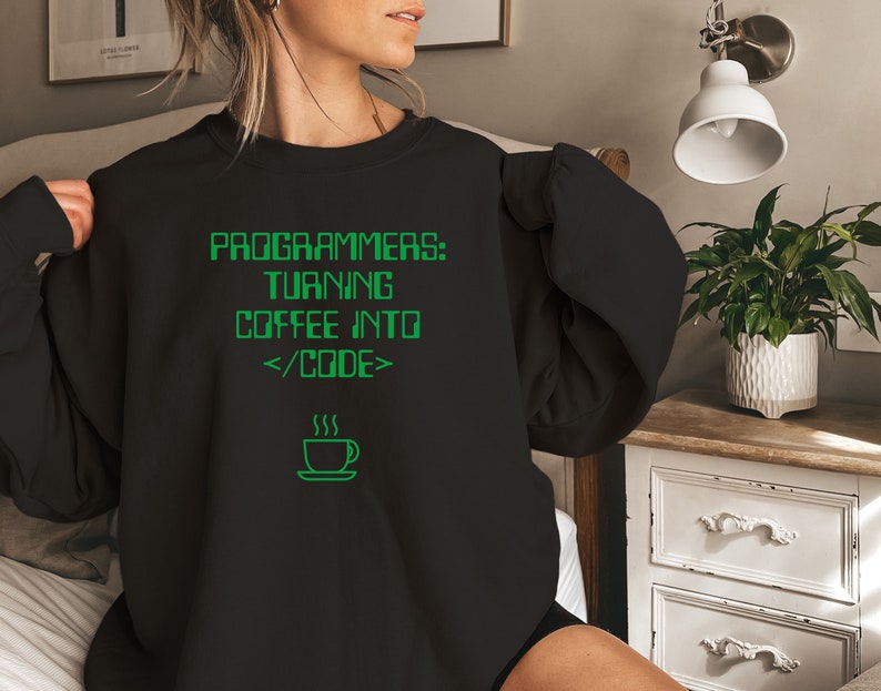 Pode incluir: Camisola preta com texto verde: "PROGRAMMERS: TURNING COFFEE INTO </CODE>". Abaixo, um gr&aacute;fico verde de uma ch&aacute;vena de caf&eacute;. A camisola &eacute; de gola redonda. O texto e o gr&aacute;fico est&atilde;o numa fonte digital.
