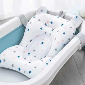 May include: A white baby bath support with a star pattern in blue, pink, and grey. The support is designed to fit inside a baby bathtub and has grey straps for securing it. The support has a headrest with two small ear-shaped extensions.