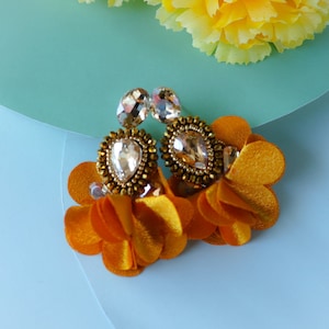 Puede incluir: Un par de pendientes de tono dorado con piedras de cristal transparentes y detalles de flores de tela naranja.