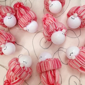 May include: A string of pink and white yarn lights shaped like little hats with white light bulbs inside. Each light bulb has a simple smiley face drawn on it.
