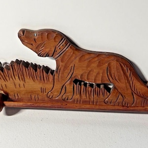 May include: A carved wooden dog wall hanging. The dog is depicted in a walking pose, with detailed fur and a collar. The base features carved grass and two small dowels for hanging. The wood has a warm, natural finish.