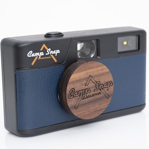 May include: A black and blue camera with a wooden lens cap. The camera has the words "Camp Snap" printed on the front and lens cap. The camera has a flash and a button on top.