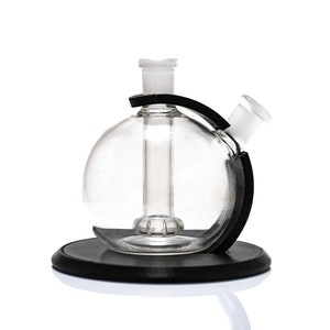 Puede incluir: Un bong de vidrio transparente con una cámara redonda y un tallo lateral, sostenido por una base negra y un soporte negro. El bong está diseñado para fumar y tiene una estética moderna y minimalista.