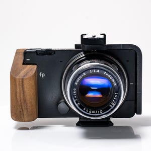 May include: Black camera with a wooden grip and a silver lens. The lens has the text "Auto-S 1:1.4 f=40mm" and "Olympus". The camera body has the letters "fp".