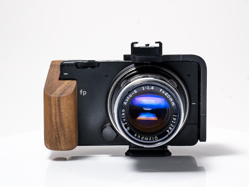 May include: Black camera with a wooden grip and a silver lens. The lens has the text "Auto-S 1:1.4 f=40mm" and "Olympus". The camera body has the letters "fp".