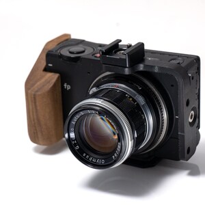 May include: Black digital camera with a wooden grip and a silver and black lens. The lens has the text "G. Zuiko Auto-S 1:1.4" and "Olympus" printed on it. The camera has the text "fp" printed on the side.