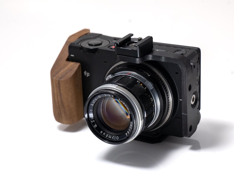 May include: Black digital camera with a wooden grip and a silver and black lens. The lens has the text "G. Zuiko Auto-S 1:1.4" and "Olympus" printed on it. The camera has the text "fp" printed on the side.