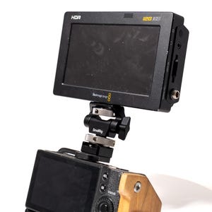 May include: A black HDR video monitor with the text "12G SDI" and "Blackmagic Design 8" is mounted on a camera via a SmallRig bracket. The camera has a wooden grip. The monitor is angled towards the viewer.