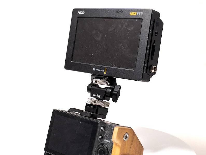 May include: A black HDR video monitor with the text "12G SDI" and "Blackmagic Design 8" is mounted on a camera via a SmallRig bracket. The camera has a wooden grip. The monitor is angled towards the viewer.
