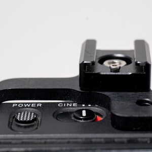 May include: Close-up of a black camera accessory with a top-mounted shoe and control buttons. The text "POWER" and "CINE" are visible. The accessory is made of metal and plastic.