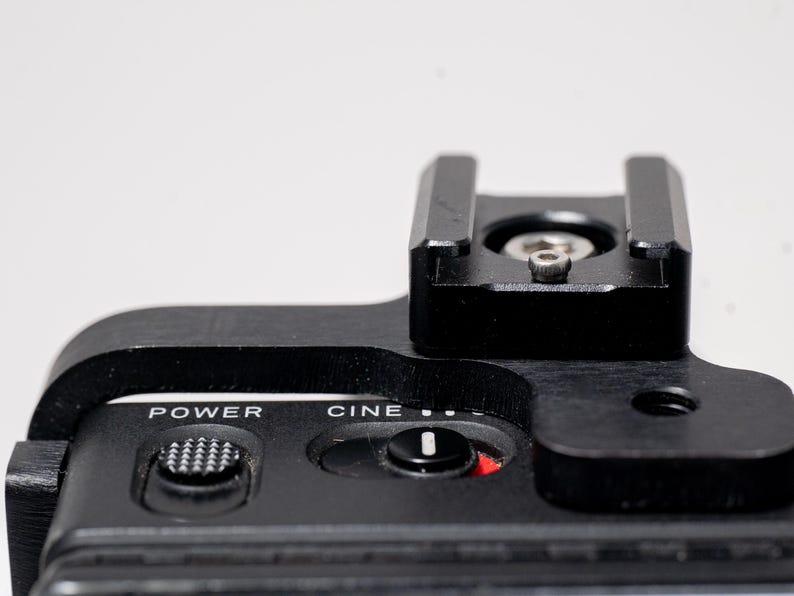 May include: Close-up of a black camera accessory with a top-mounted shoe and control buttons. The text "POWER" and "CINE" are visible. The accessory is made of metal and plastic.