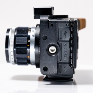 May include: A black camera with a silver lens, featuring a black lens ring with white numbers and markings. The camera body has a black exterior with ports labeled HDMI and MIC. A small black accessory is mounted on top.