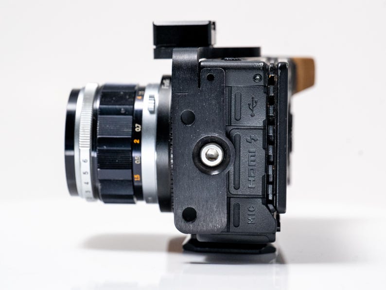 May include: A black camera with a silver lens, featuring a black lens ring with white numbers and markings. The camera body has a black exterior with ports labeled HDMI and MIC. A small black accessory is mounted on top.