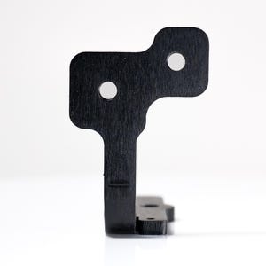 May include: A black, geometric metal bracket with two circular holes and a textured surface. The bracket has a unique shape with a rectangular base and a top section with rounded corners. The object is set against a white background.