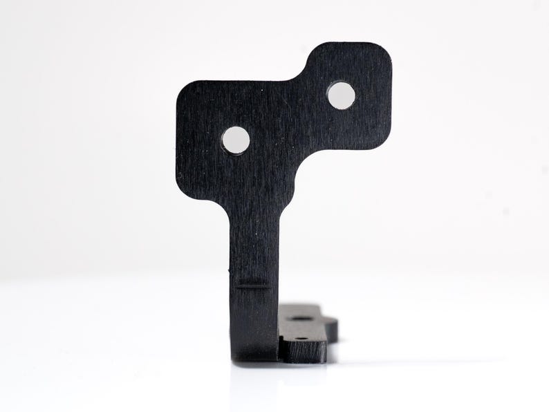 May include: A black, geometric metal bracket with two circular holes and a textured surface. The bracket has a unique shape with a rectangular base and a top section with rounded corners. The object is set against a white background.