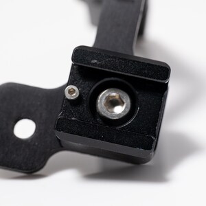 May include: Close-up of a black metal component with a square base and a circular hole. A silver bolt is centered within the square base. The component is likely part of a larger mechanical assembly.