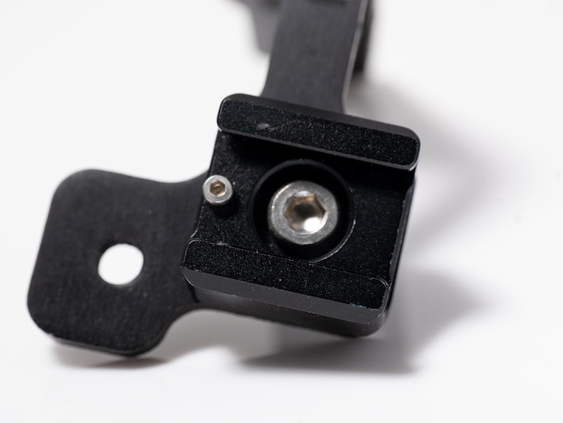 May include: Close-up of a black metal component with a square base and a circular hole. A silver bolt is centered within the square base. The component is likely part of a larger mechanical assembly.