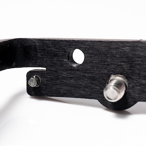May include: Close-up of a black, textured metal object with a series of holes and silver metal fasteners. The object has a dark, wood-grain appearance and is set against a white background.