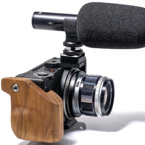 May include: Black digital camera with a lens and a wooden grip. A black microphone with a foam windscreen is mounted on top. The camera lens has measurement markings.