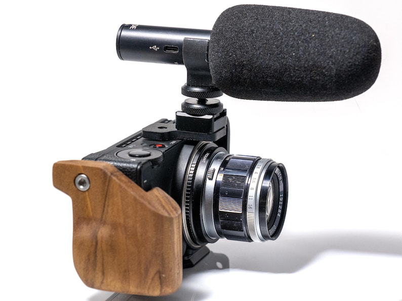 May include: Black digital camera with a lens and a wooden grip. A black microphone with a foam windscreen is mounted on top. The camera lens has measurement markings.