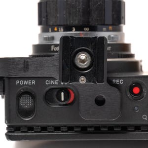 May include: Close-up of a black camera with a lens. The camera has a black top with the words "POWER", "CINE", and "REC" printed on it. A red button is visible near the "REC" label. The lens is black and silver.