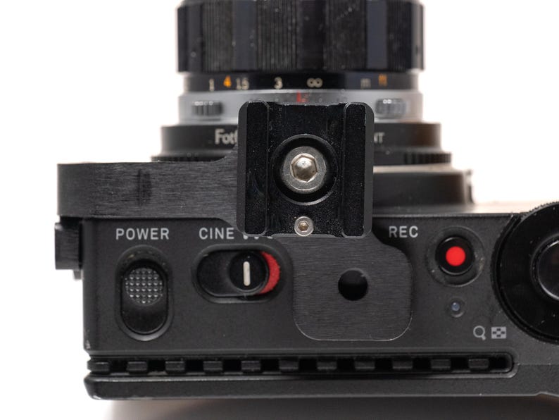 May include: Close-up of a black camera with a lens. The camera has a black top with the words "POWER", "CINE", and "REC" printed on it. A red button is visible near the "REC" label. The lens is black and silver.