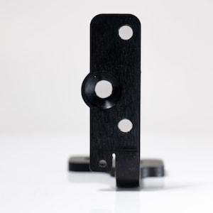 May include: A black metal bracket with a rectangular body, featuring a circular indentation and two smaller holes. The bracket has a small, rectangular base and a slot on the bottom edge.