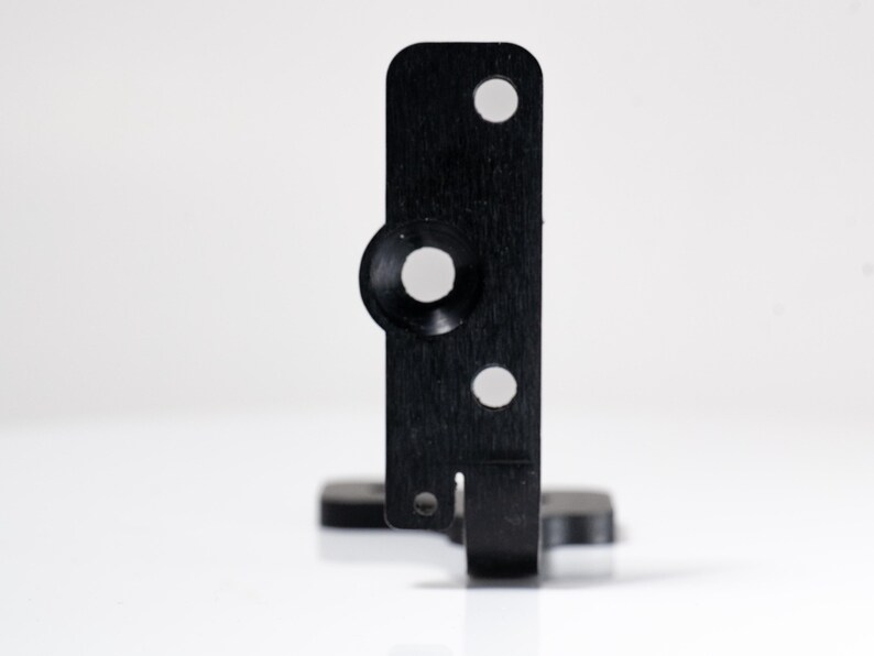 May include: A black metal bracket with a rectangular body, featuring a circular indentation and two smaller holes. The bracket has a small, rectangular base and a slot on the bottom edge.