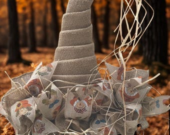 May include: A tall, conical hat crafted from tan burlap, featuring a spiraled structure. The base is adorned with loops of light-colored ribbon printed with scarecrow faces and sunflowers, accented by strands of natural raffia straw.