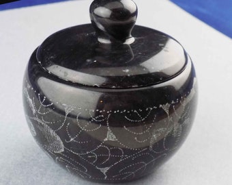 May include: A black, round, lidded container with a swirl design carved into the surface. The container is made of polished stone.
