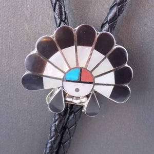 May include: A Native American-inspired bolo tie with a black, white, and red geometric design on a mother-of-pearl background. The bolo tie is made of sterling silver and has a black leather cord.