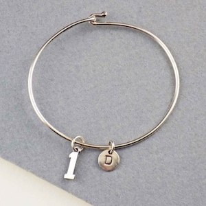 May include: A silver-colored bangle bracelet with a hook closure. The bracelet features two charms: a number 1 charm and a round charm with the letter "D". The bracelet is displayed on a gray surface.