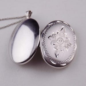 May include: An oval silver locket pendant on a thin chain. The locket is open, revealing a smooth, polished interior on the left side and an exterior on the right side featuring an engraved floral design surrounded by a decorative serrated border.