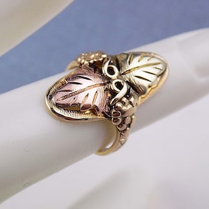 May include: A gold ring featuring a leaf and grape design. The ring has a gold band and a detailed design with gold, rose gold, and green gold leaves and grapes. The ring is set on a white surface.